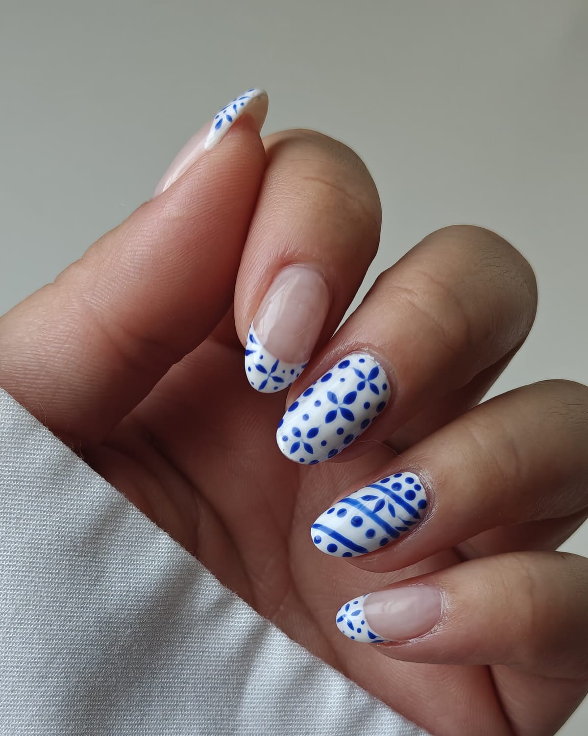 Close-up of almond nails painted with a blue and white Portuguese tile pattern over a sheer nude base, against a white cuff.