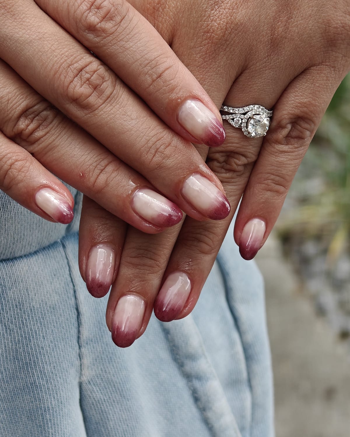Almond nails in a sheer milky base fading into deep burgundy tips, a soft ombre French, hands crossed over pale denim.