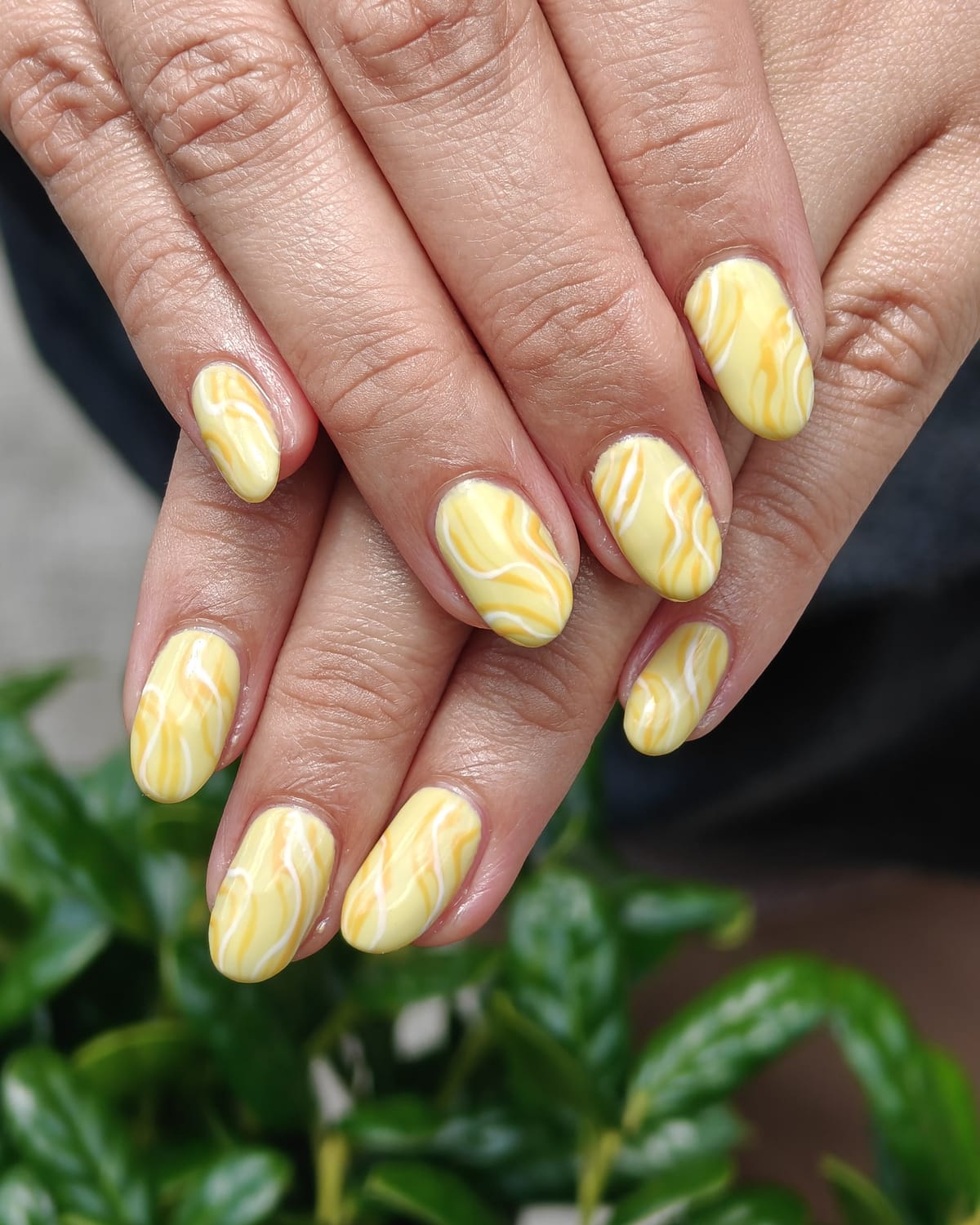 Butter-yellow almond nails with soft white marble swirls, hands resting over a green garden hedge.