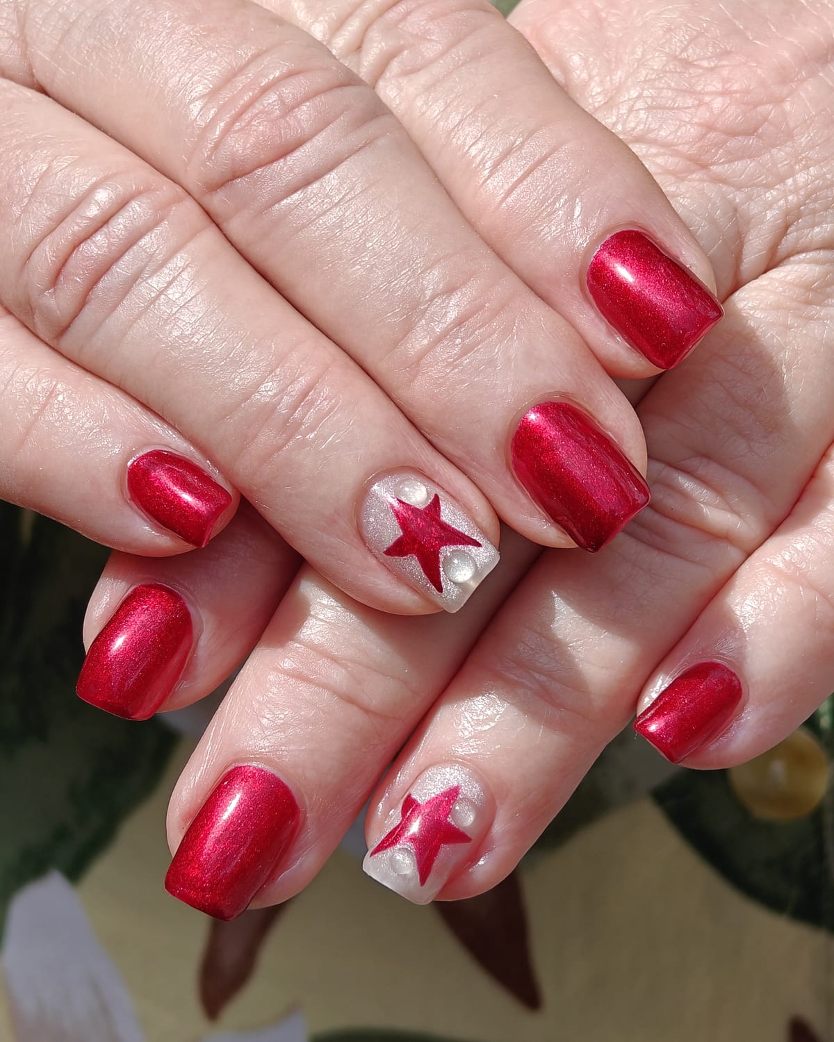 Long nails in deep cherry red metallic with two white accent nails carrying hand-painted red stars, hands crossed.