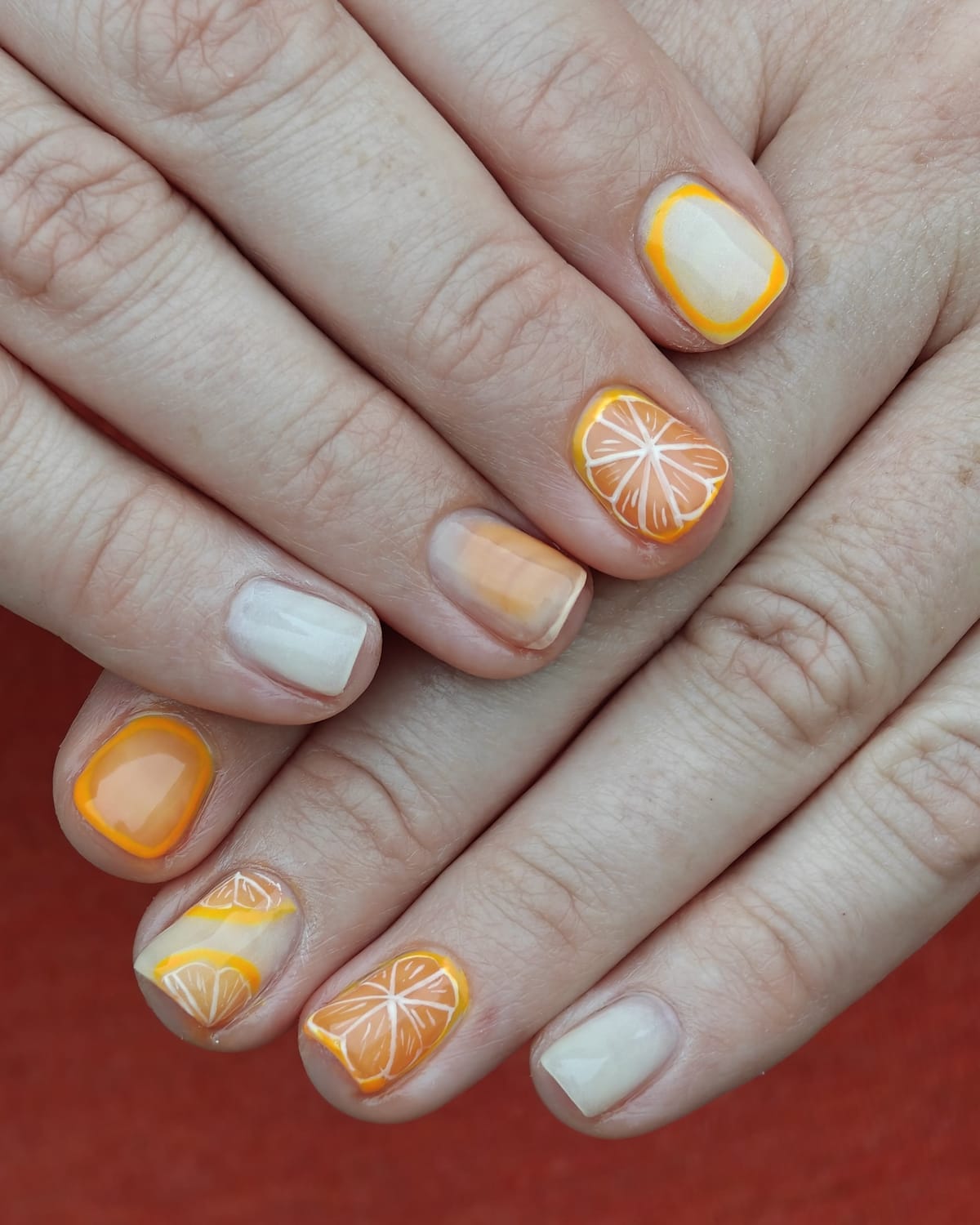 Short nails in cream and marigold orange with hand-painted orange and lemon slices, hands crossed on a terracotta background.