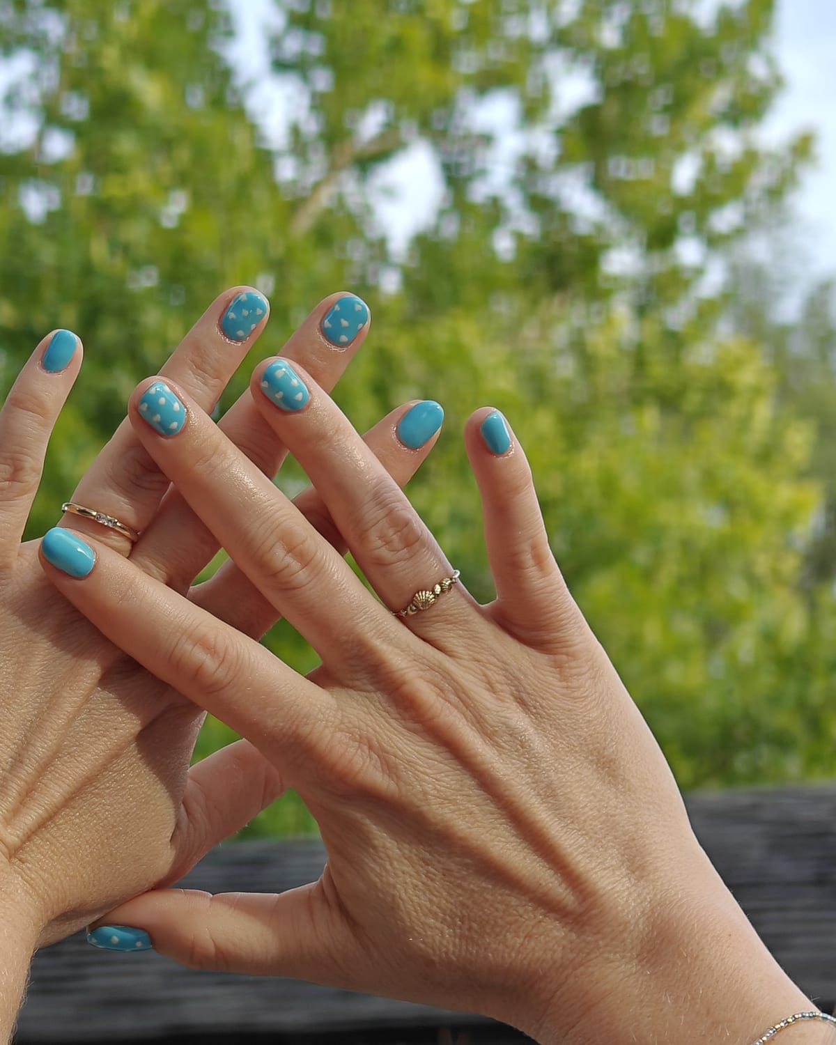 Sky blue nails with soft white cloud art and a turquoise shimmer accent, fingers spread against real sky and treetops.