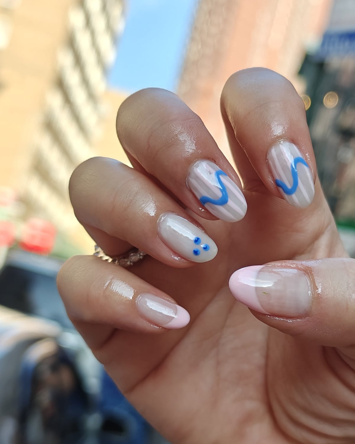 Long almond nails in sheer nude with cobalt blue squiggle and dot line art on the accent nails, hand raised on a city street.