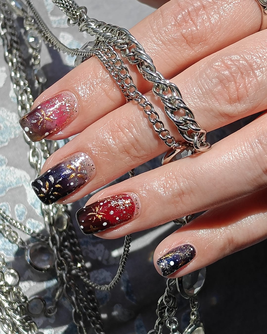 Long square nails in black, burgundy and charcoal glitter with gold and silver firework bursts hand-painted across the tips.