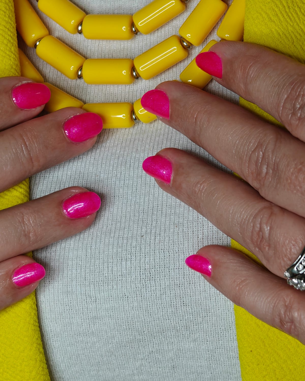 Short nails in vivid fuchsia with a high-gloss finish, hands framing a chunky mustard yellow beaded necklace over a cream knit.