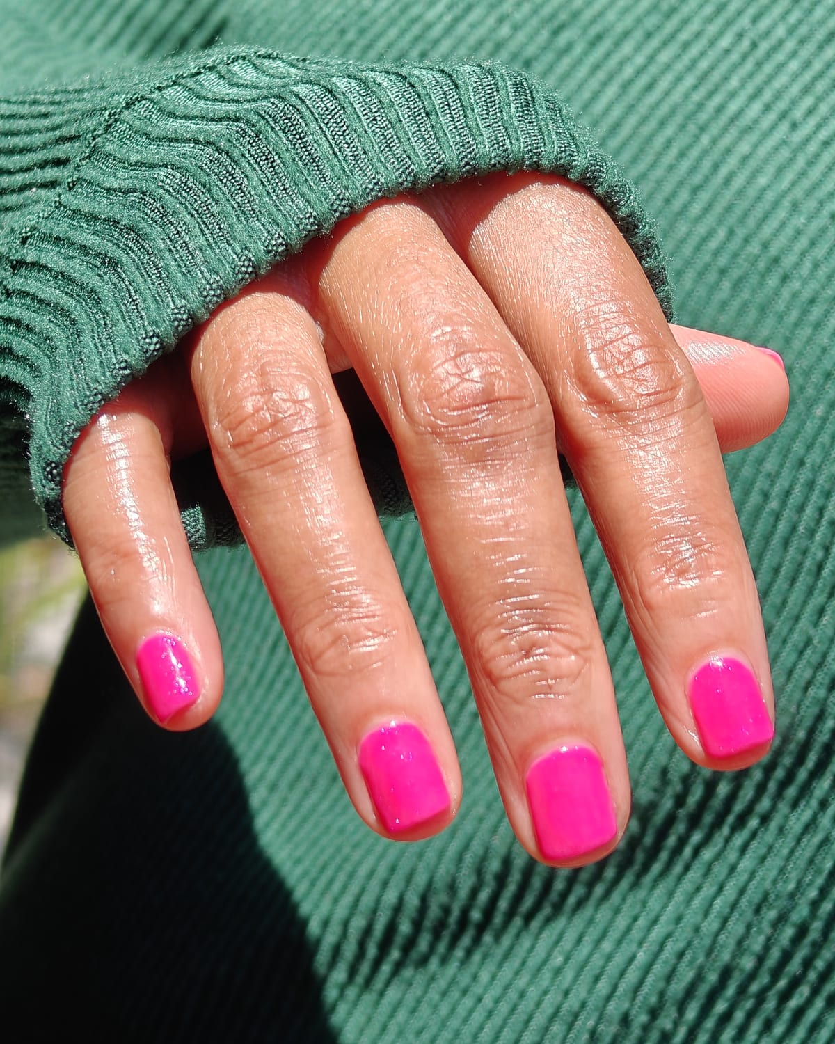 Short square nails in bright fuchsia with a glossy finish, one hand resting against the ribbed cuff of a green sweater.
