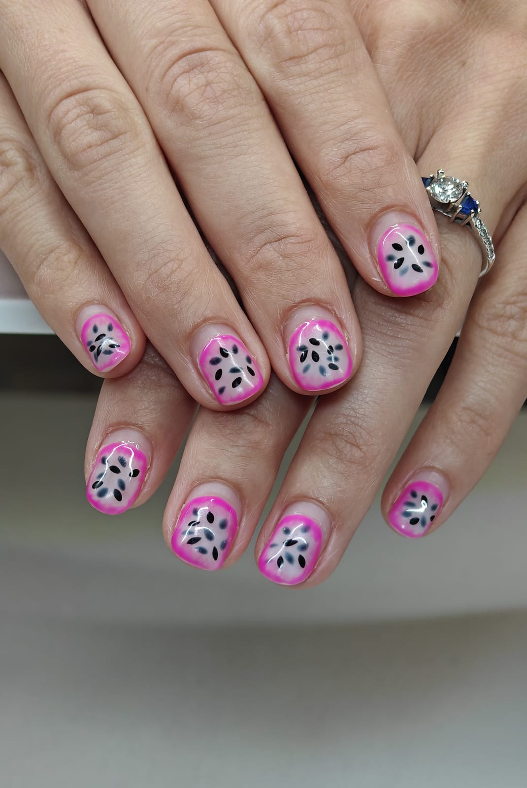 Short nails with hot pink French tips over a white base, speckled with painted black seeds in a dragon fruit pattern.