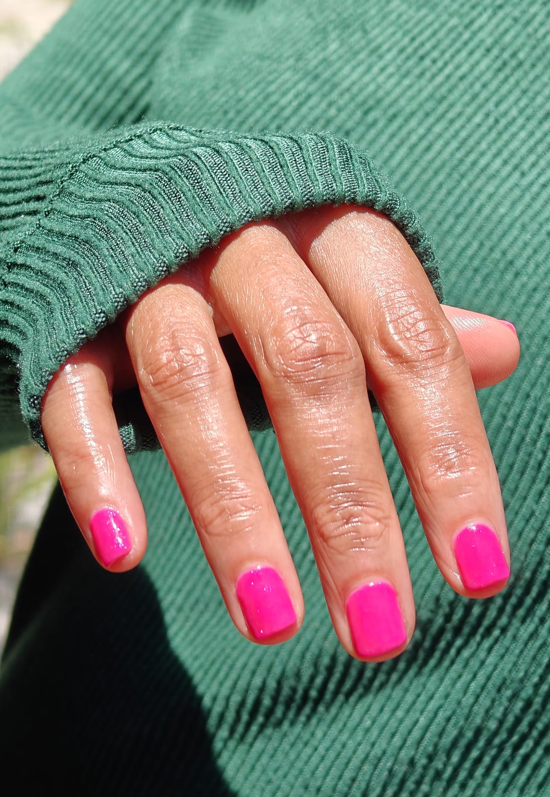 Short square nails in bright fuchsia with a glossy finish, one hand resting against the ribbed cuff of a green sweater.