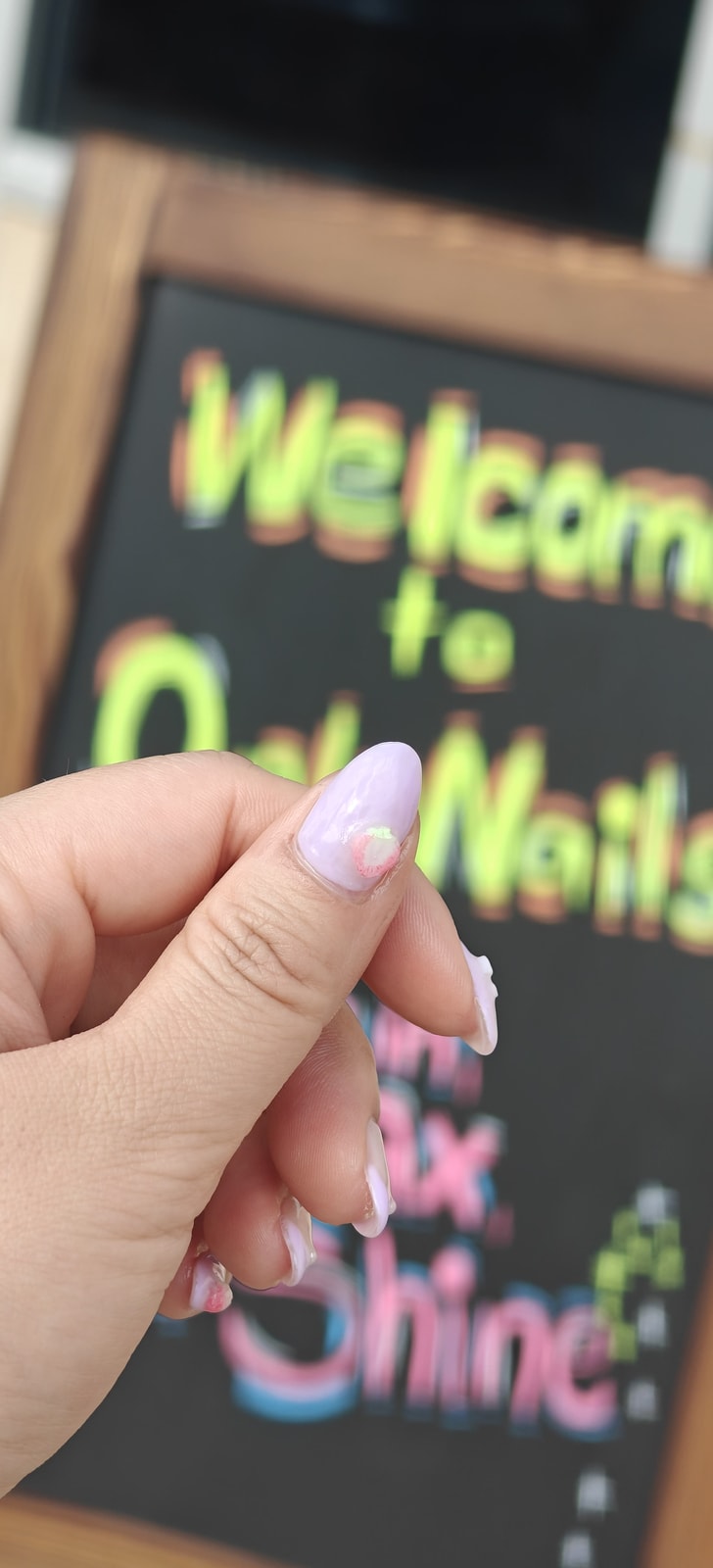 A single lilac marbled almond nail held up in front of the handwritten Only Nails welcome chalkboard.