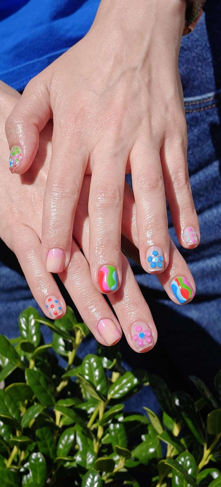 Short nails with neon confetti dot art in pink, blue and green over a sheer pink base, hands in bright sunlight above a hedge.
