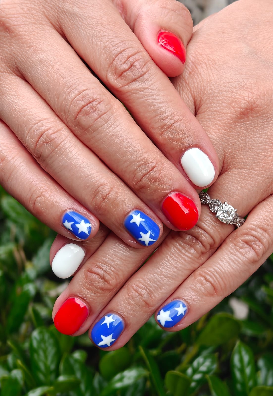 Short nails in red, white and navy blue with crisp hand-painted white stars, hands crossed above a green hedge.