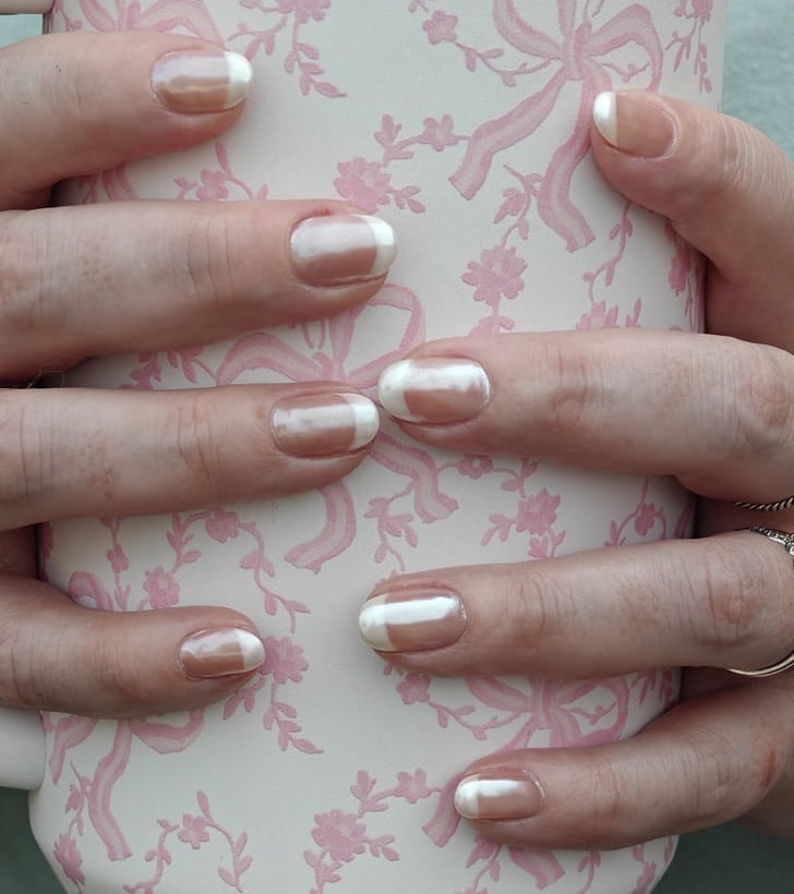Pearl chrome French nails with milky tips, hands wrapped around a cream tumbler printed with pink bows and flowers.