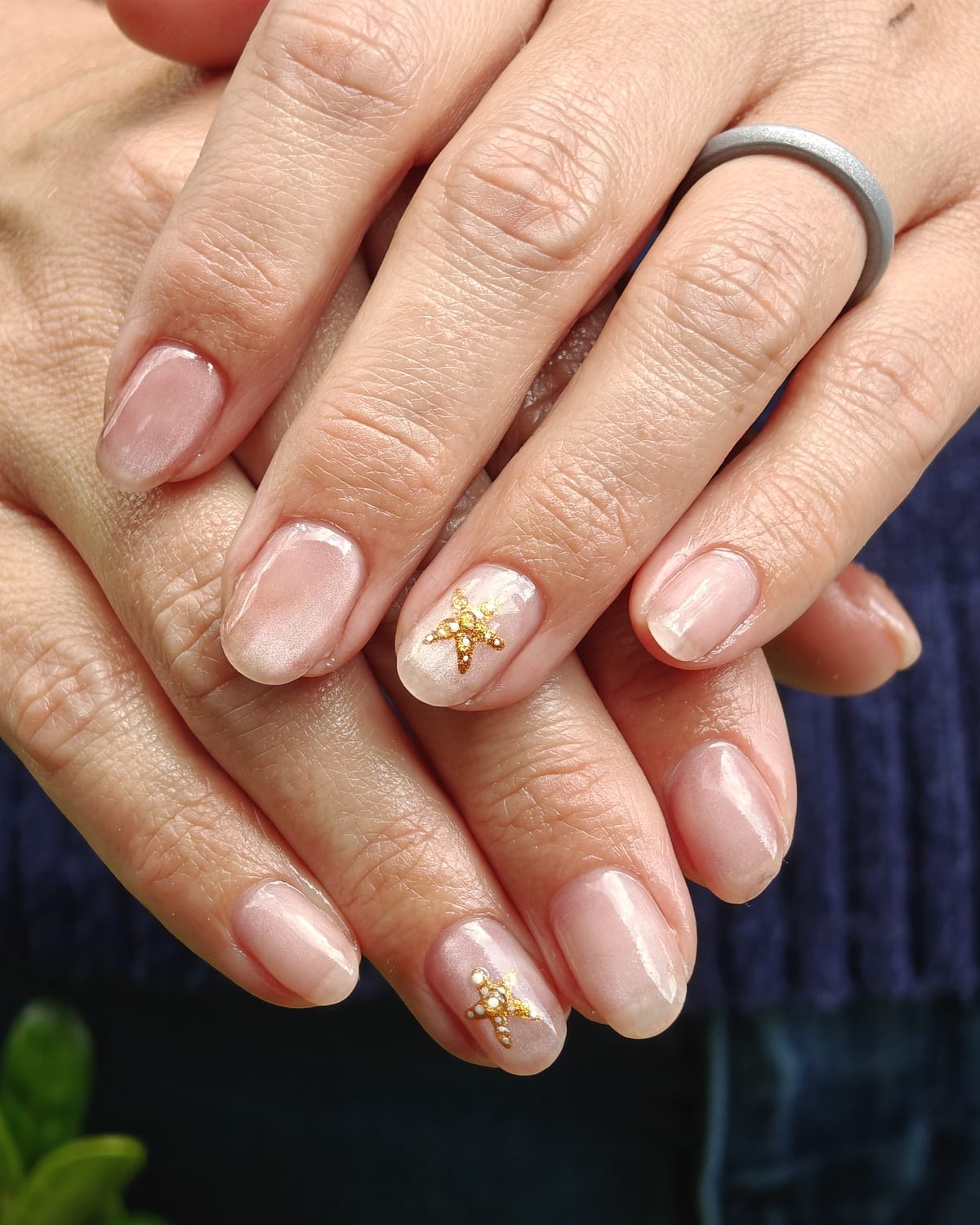 Short natural nails in a sheer blush base with gold caviar bead clusters at the cuticle, hands crossed over a navy knit.