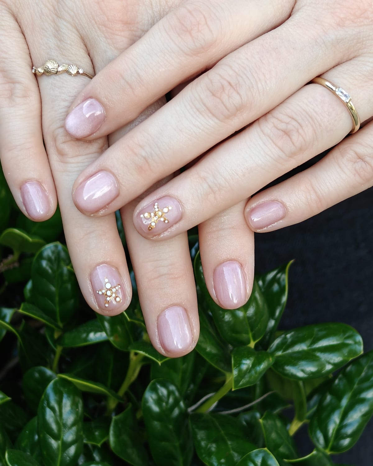 Short natural nails in a sheer blush base scattered with gold leaf flakes, hands resting on glossy green foliage.