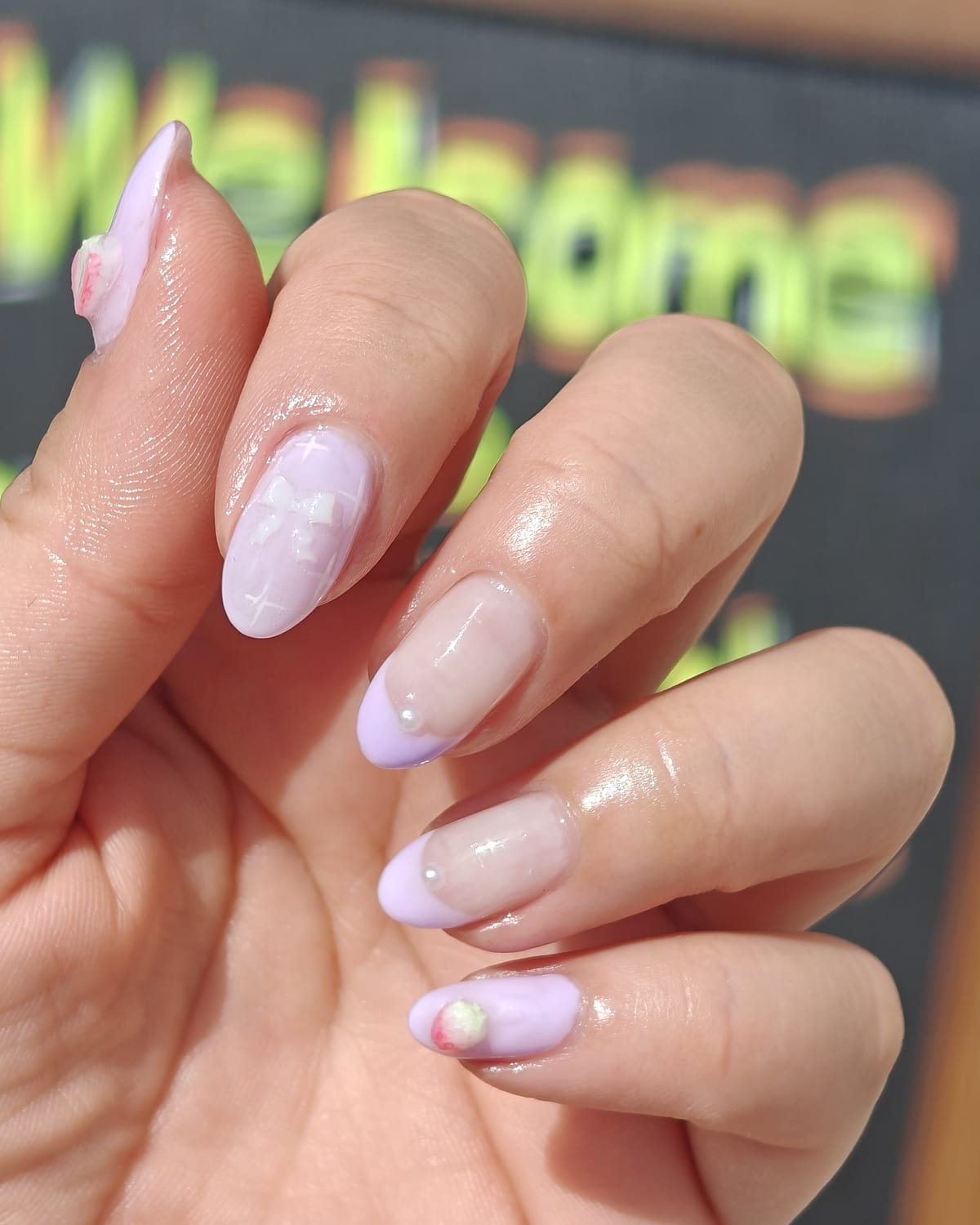 Almond nails in pastel lilac and sheer milky pink with pearl accents and a hand-painted strawberry, in front of the salon welcome sign.