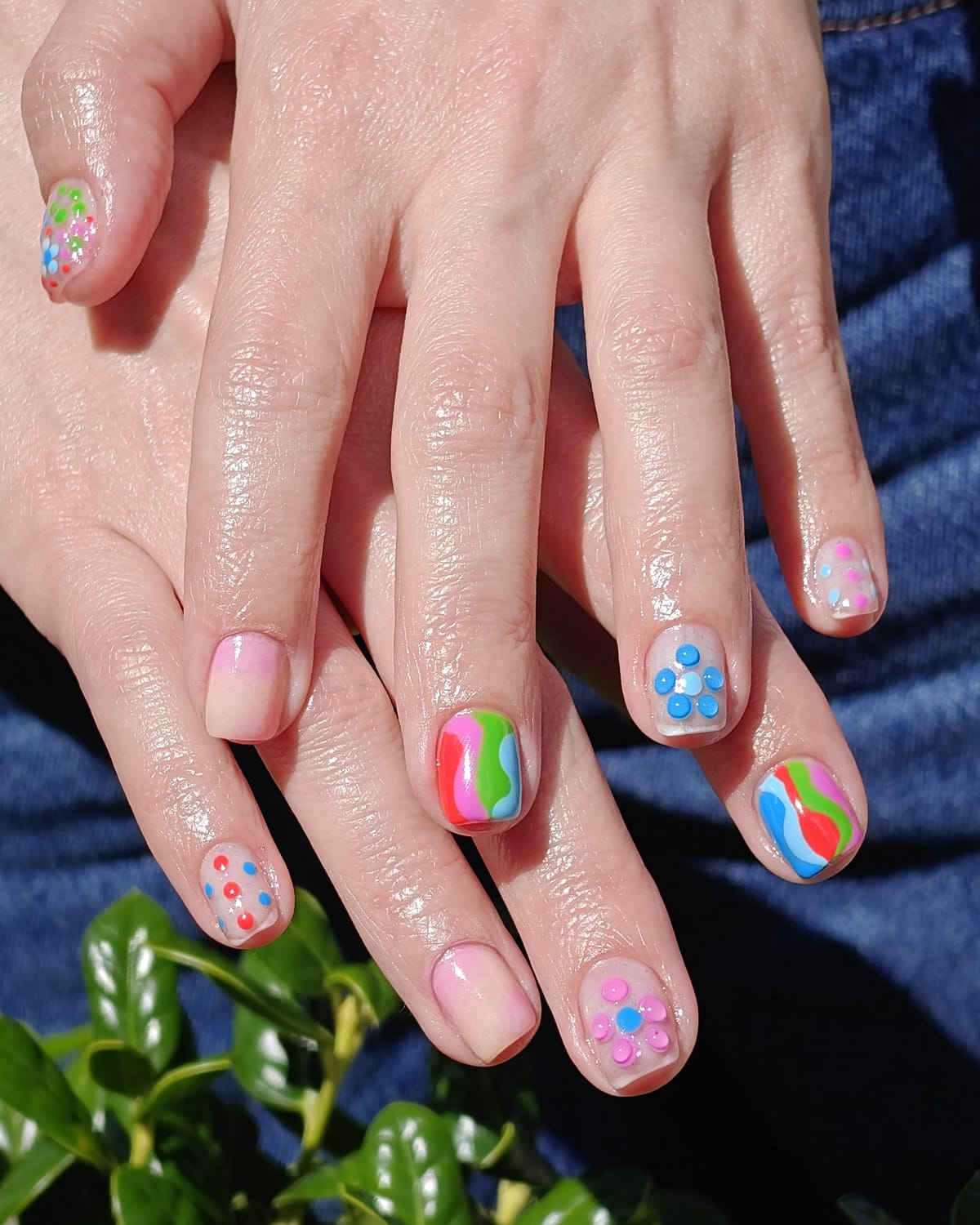 Short nails with neon confetti dot art in pink, blue and green over a sheer pink base, hands in bright sunlight above a hedge.