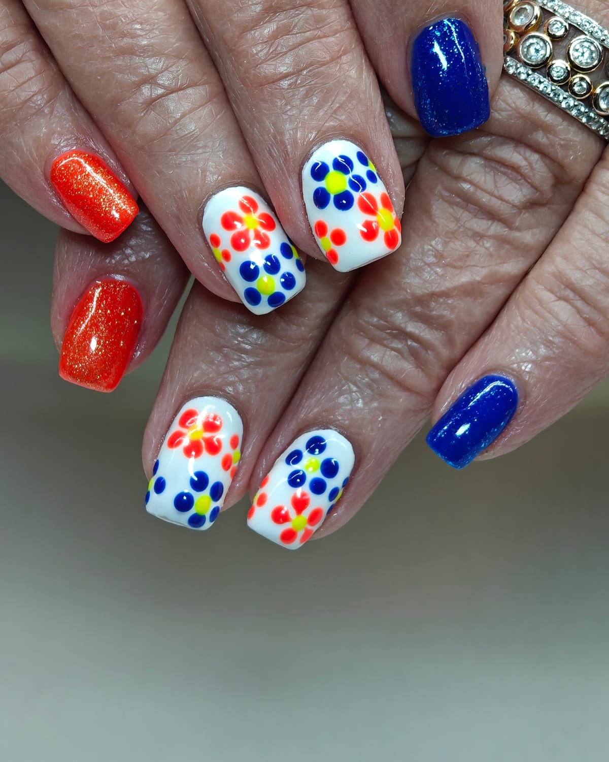 Short square nails alternating orange glitter, cobalt blue and white with hand-painted neon dot flowers in red, blue and yellow.