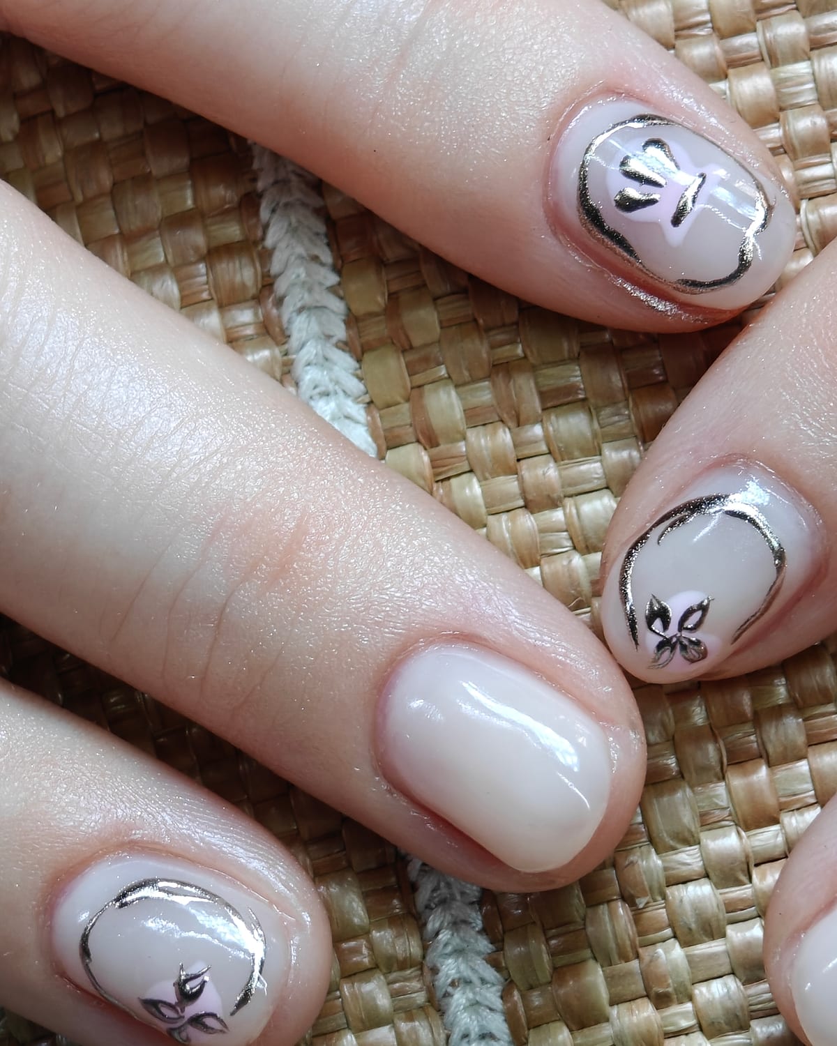 Long almond nails in a soft milky nude with delicate gold-outlined floral line art on the accent nails, hand on woven straw.