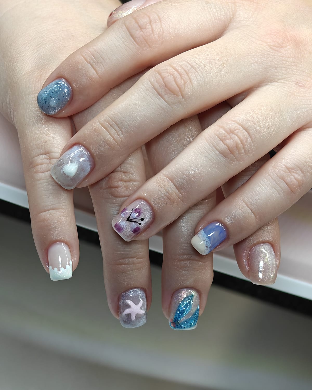 Short nails in aqua and lavender glitter with sculpted white starfish and shell accents over a sheer base, hands crossed.