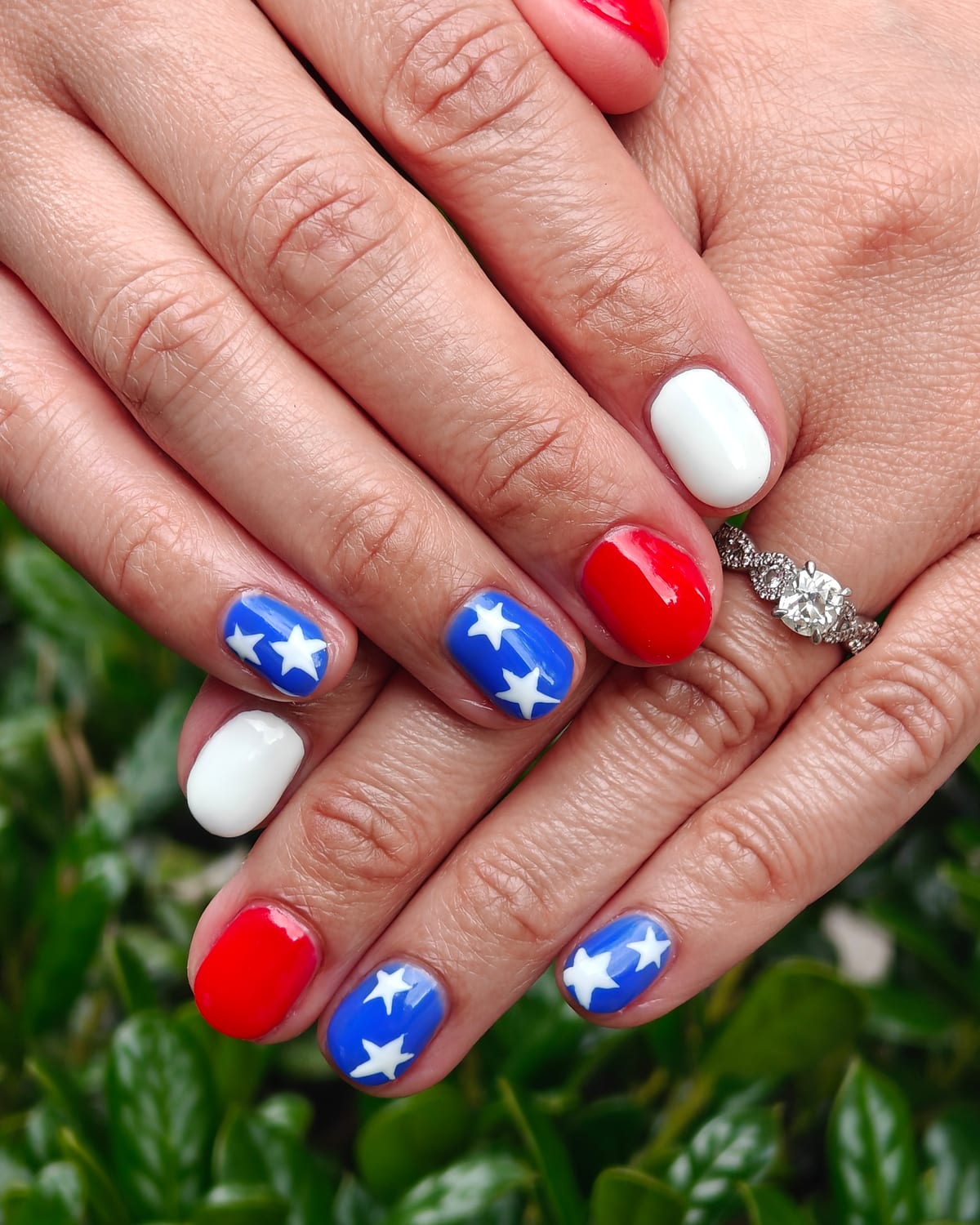 Short nails in red, white and navy blue with crisp hand-painted white stars, hands crossed above a green hedge.