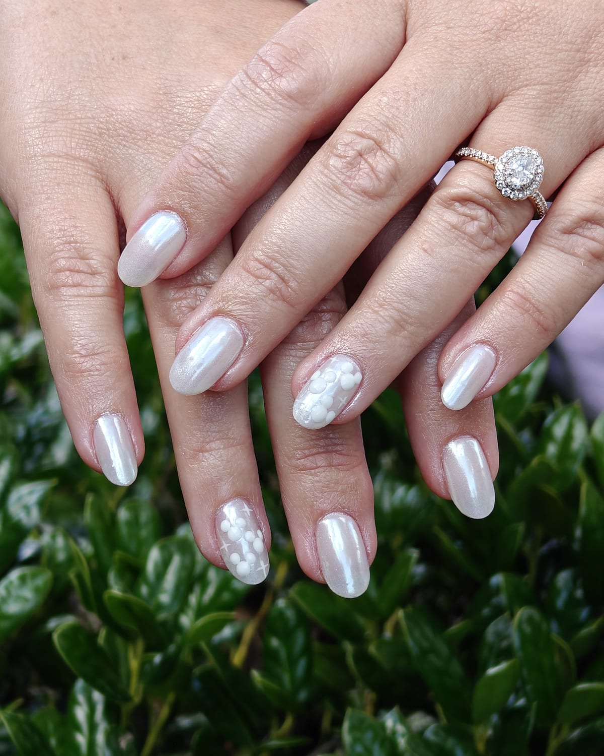 Long almond nails in pearl chrome with a soft white marble accent and delicate 3D floral detail, hands over a green hedge.