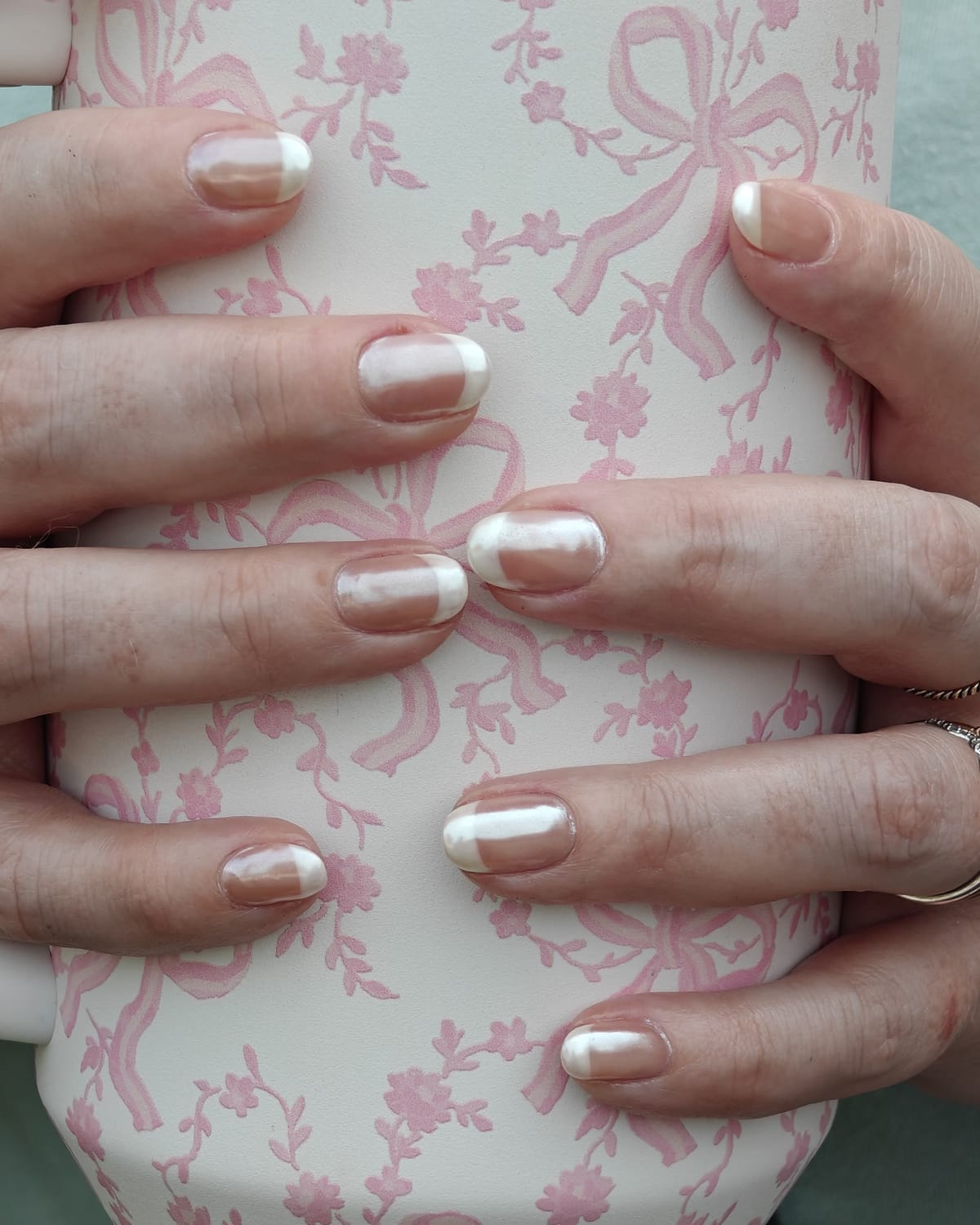 Pearl chrome French nails with milky tips, hands wrapped around a cream tumbler printed with pink bows and flowers.