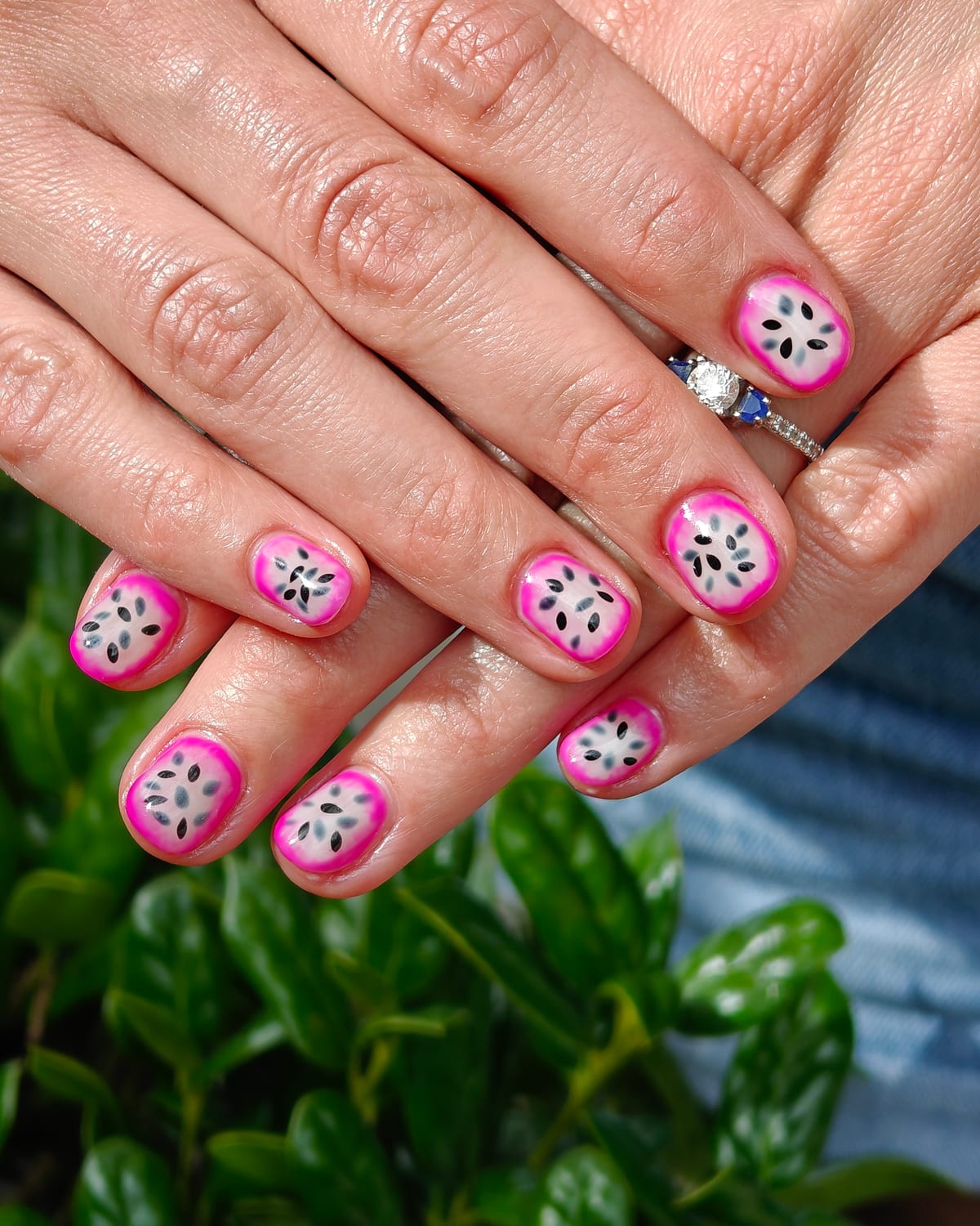 Short square nails outlined in hot pink with white centres and painted black seeds, a dragon fruit motif, hands crossed over a hedge.
