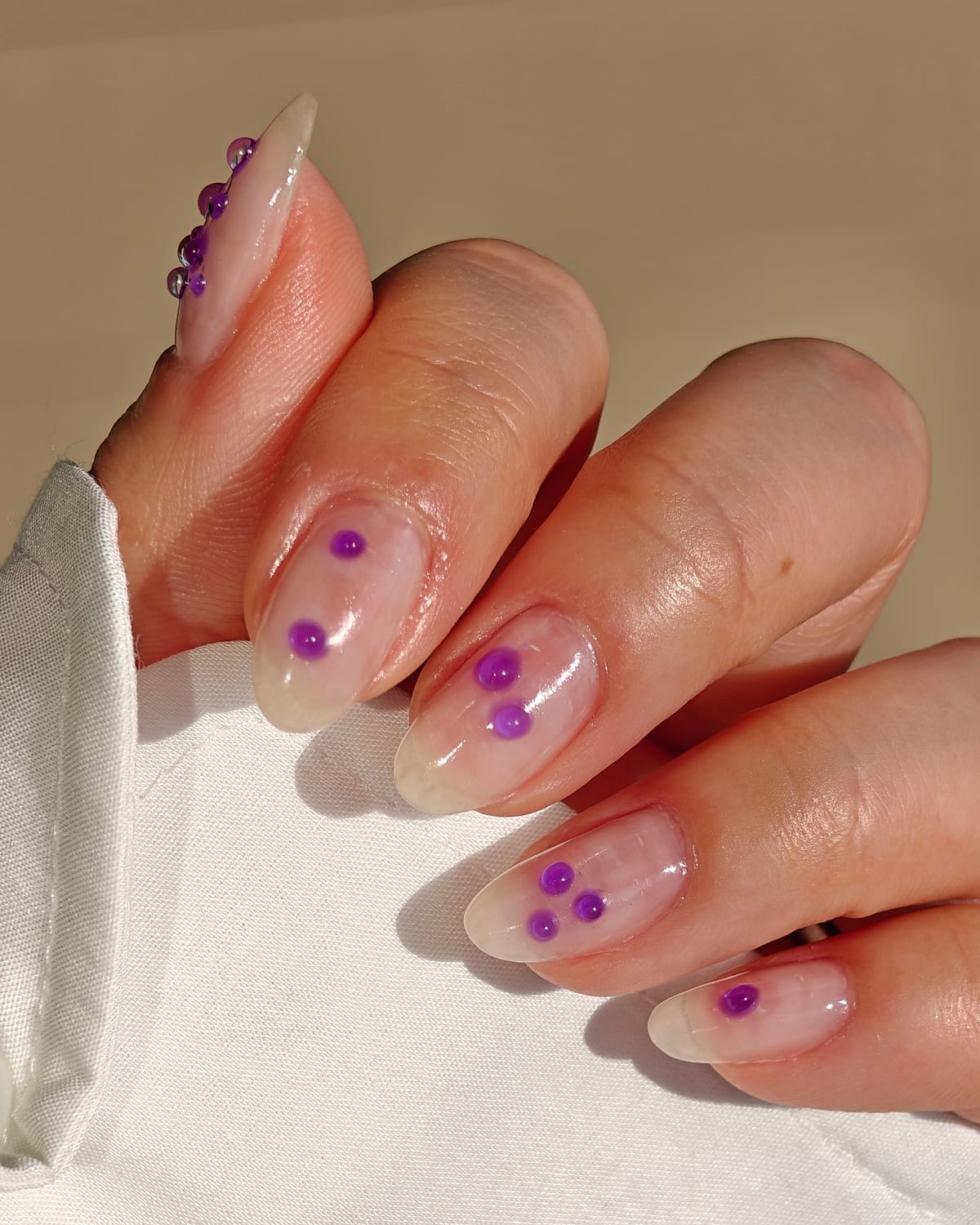 Long almond nails in sheer nude with clusters of purple caviar beads scattered across the tips, hand resting on a cream cushion.