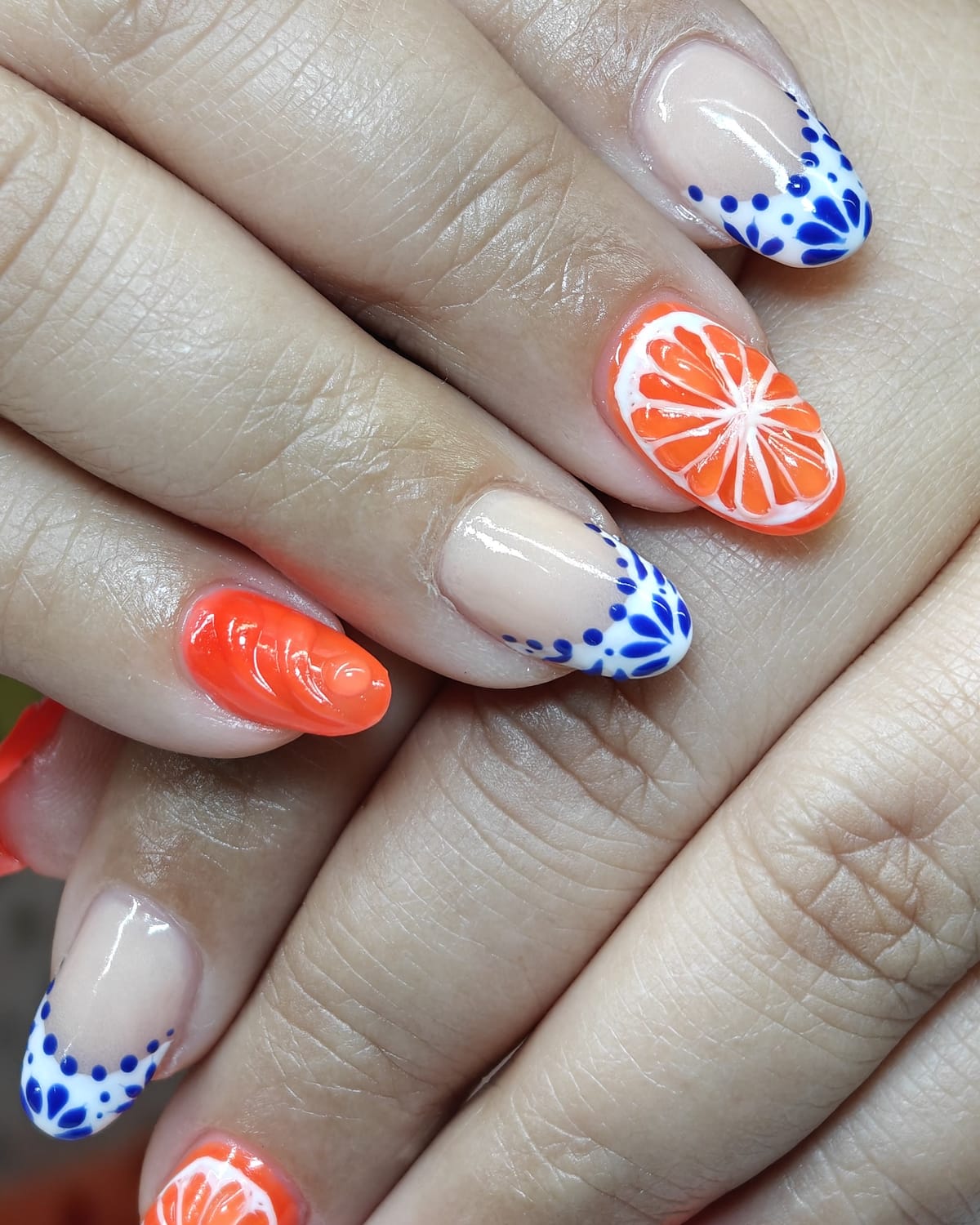 Almond nails mixing bright orange polish with blue and white Talavera tile French tips and a painted orange slice accent.