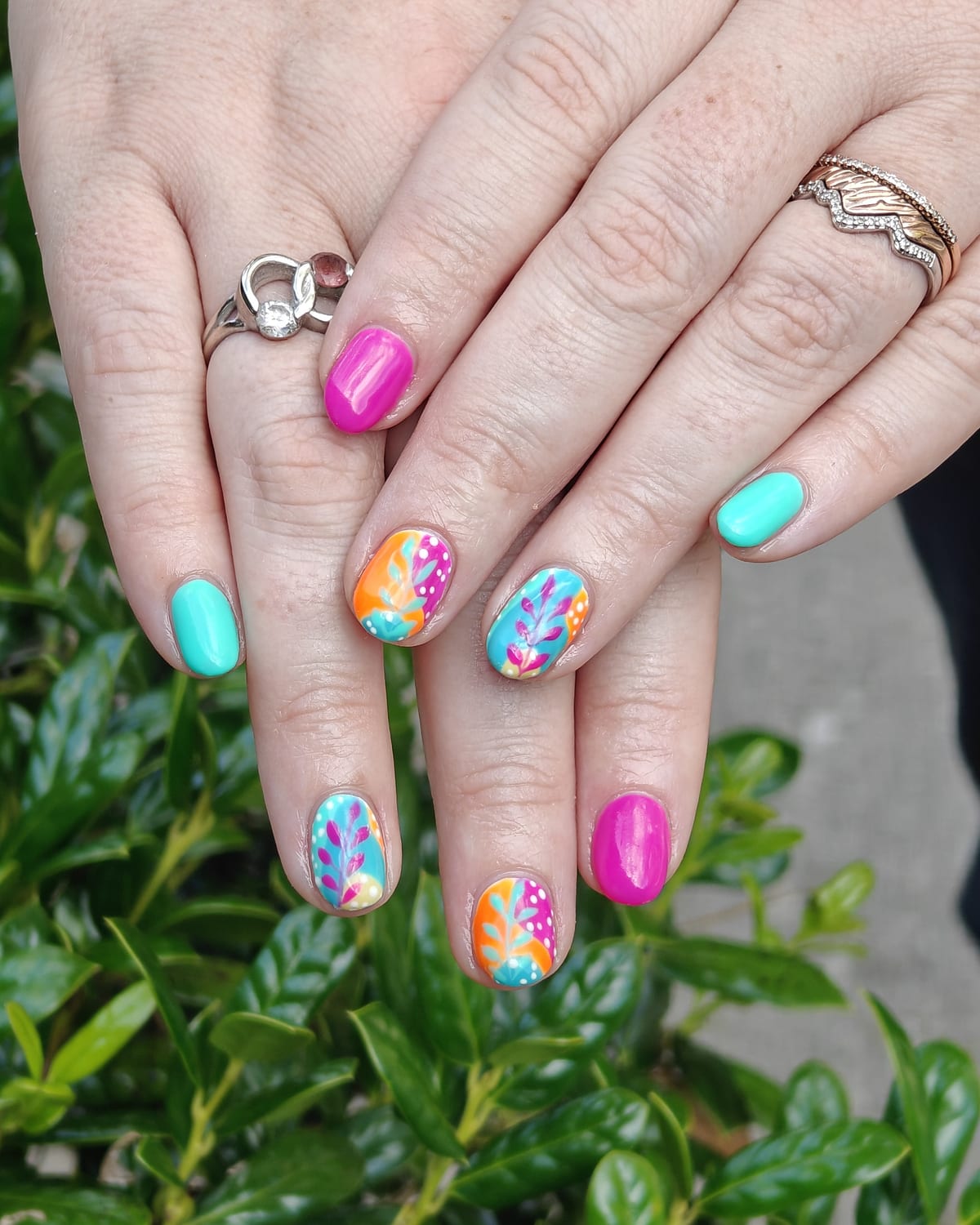 Bright turquoise, magenta and orange nails with hand-painted tropical leaves and floral accents, hands resting on a green hedge.