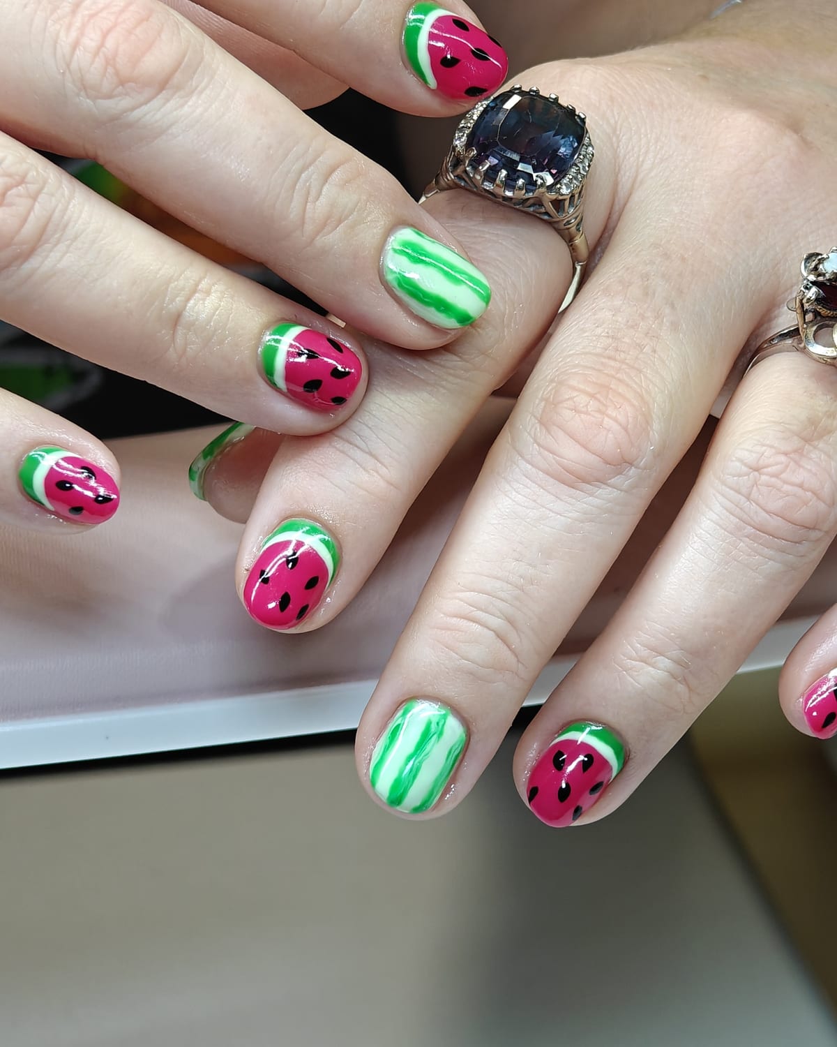 Almond nails alternating watermelon pink with black seeds and green-and-white rind stripes, hands crossed on a pale table.